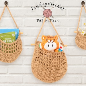 May include: Three hanging crochet baskets in varying sizes, suspended from hooks on a white brick wall. The baskets are filled with items, including a plush tiger toy, a package, and flowers. The text "Taghoycrochet Pdf Pattern" is at the top.