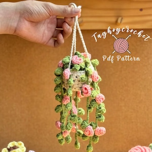May include: A handmade crochet hanging planter with pink and green floral accents. The planter is suspended by a white cord and features the text "Taghoycrochet Pdf Pattern". The crochet work is detailed and the colors are vibrant.