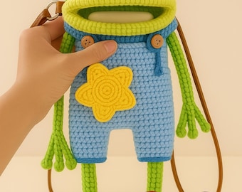 May include: A handmade crochet frog-shaped crossbody bag. The bag features a green head with large black and white eyes, pink cheeks, and a yellow mouth. The frog wears blue overalls with a yellow star and brown buttons. The bag has a brown strap.