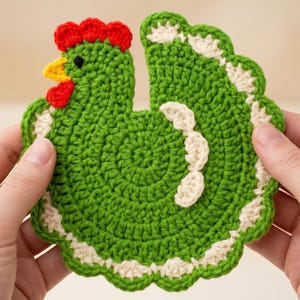 May include: A green crocheted coaster shaped like a chicken, featuring a bright red comb, a yellow beak, and a small black eye. The edges of the chicken are accented with a scalloped white border, and a white curved detail is stitched on the body.