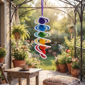 May include: A hanging crochet spiral mobile featuring six spheres in a vertical rainbow gradient of purple, blue, green, yellow, orange, and red. Each sphere is encircled by a white crochet ring, suspended by a purple cord from a metal frame.