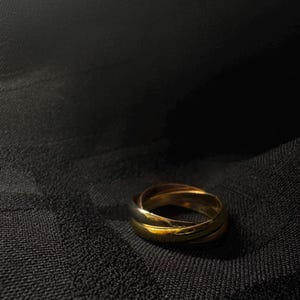 May include: A gold ring with three intertwined bands, set against a dark, textured background. The ring's polished surface reflects light, highlighting its design. The jewelry piece is a classic accessory.
