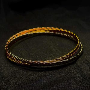 May include: A gold-toned, braided bracelet with a textured design. The bracelet features a mix of gold, copper, and black strands, creating a multi-coloured, metallic appearance. The jewellery is set against a dark background.