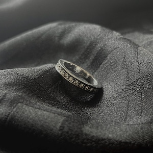 May include: A silver ring with a band of small, clear gemstones. The ring is set against a dark, textured background, creating a contrast that highlights the ring's sparkle. The ring appears to be a wedding band or eternity ring.
