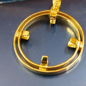 Puede incluir: Un colgante dorado con un diseño circular. El colgante tiene una fianza texturizada y tres cierres espaciados uniformemente alrededor del círculo interior. El colgante está sobre un fondo azul.