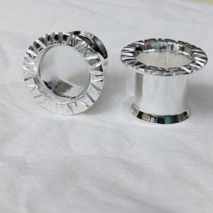 Puede incluir: Dos expansores de oreja plateados. Uno está tumbado de lado, revelando un centro hueco. El otro está en posición vertical. Ambos tienen un borde decorativo y estriado. Los expansores están sobre una superficie blanca.