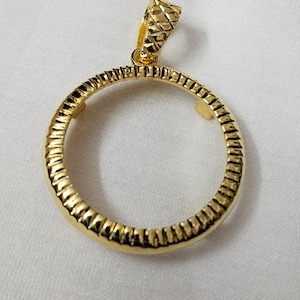 May include: A gold-coloured pendant with a textured, circular design. The pendant has a textured bail for attaching to a chain. The circular portion of the pendant has a ribbed pattern around the outer edge.