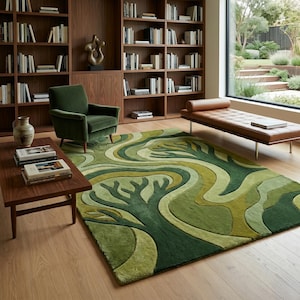 May include: A green area rug with a tree design in varying shades of green. The rug is placed in a living room with a green armchair, a wooden coffee table, and a brown leather chaise lounge. Bookshelves are in the background.