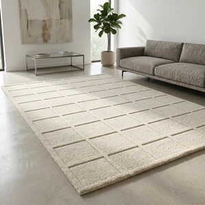 May include: A large, rectangular, cream-coloured rug with a geometric grid pattern. The rug is made of a soft, textured material and is placed on a light-coloured concrete floor. The rug is in a modern living room setting.