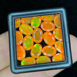 May include: A selection of oval-shaped, bright orange opals with green and yellow highlights, presented in a black square frame. The gemstones are closely packed, highlighting their polished surfaces and internal colour variations. The opals are set against a dark backdrop.