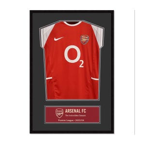 Puede incluir: Camiseta de fútbol del Arsenal FC enmarcada, roja con detalles blancos, con el logo O2 y el escudo del Arsenal. El marco negro rodea la camiseta y una placa en la parte inferior dice "Arsenal FC The Invincibles Season Premier League 2003/04."