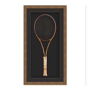 May include: A framed vintage tennis racket. The racket has a gold-colored frame and a black stringed head. The handle is wrapped in a light-colored grip. The frame is a dark brown color.