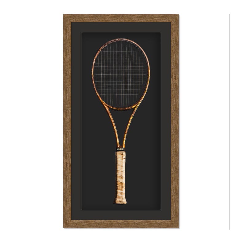 May include: A framed vintage tennis racket. The racket has a gold-colored frame and a black stringed head. The handle is wrapped in a light-colored grip. The frame is a dark brown color.