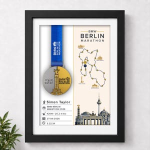 Pode incluir: Um quadro com uma medalha da Maratona de Berlim BMW numa fita azul, um mapa do percurso e uma placa personalizada para Simon Taylor com a data 27-09-2026, 42 km e um tempo de 3.22.34, ao lado de ilustrações de monumentos de Berlim.