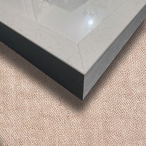 May include: A close-up of a picture frame with a light gray wooden frame and a black interior. The frame has a glass front and is set against a textured, tan background.