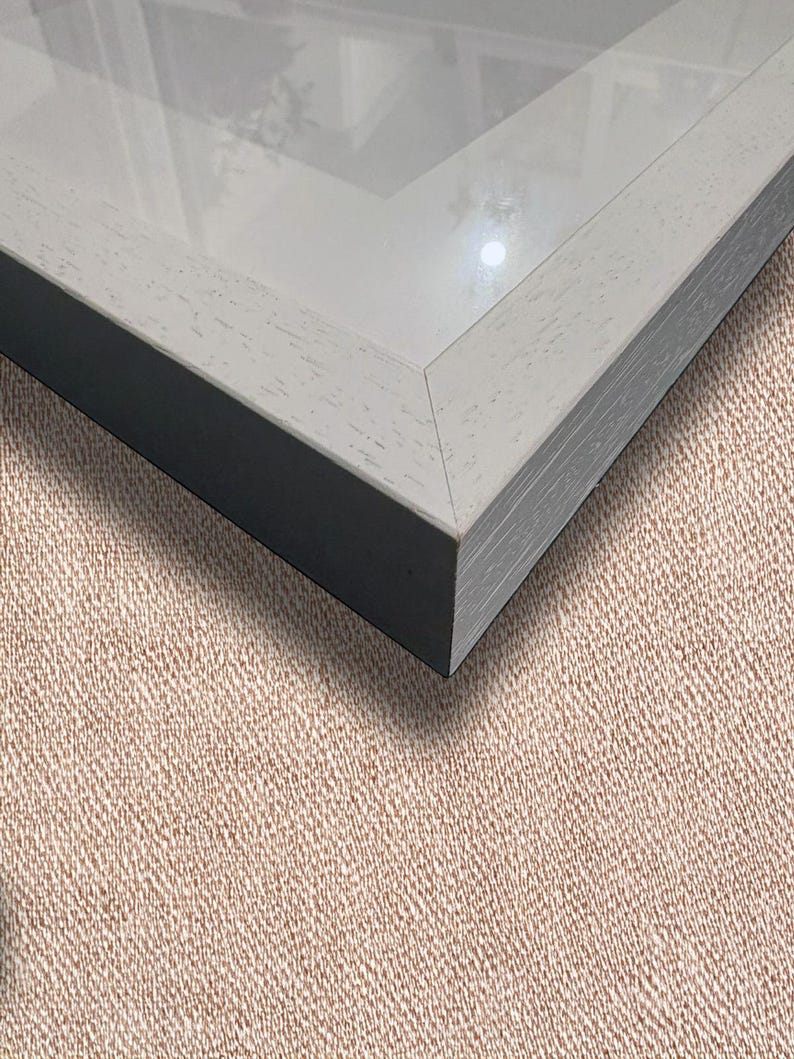 May include: A close-up of a picture frame with a light gray wooden frame and a black interior. The frame has a glass front and is set against a textured, tan background.