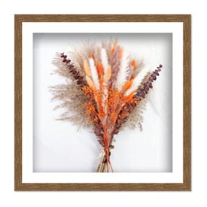 May include: A framed floral arrangement featuring a variety of dried flowers in shades of orange, white, and brown. The bouquet is tied with twine and set against a white background. The frame is a light brown color.
