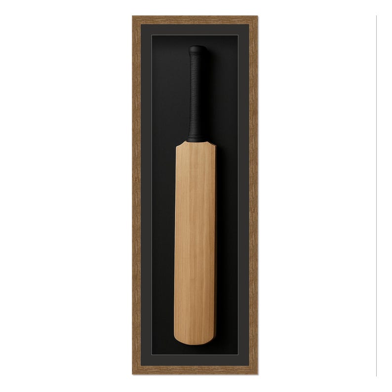 May include: A framed cricket bat. The bat is made of light-colored wood with a black grip. It is set against a black background within a brown wooden frame. The frame is rectangular and vertically oriented.