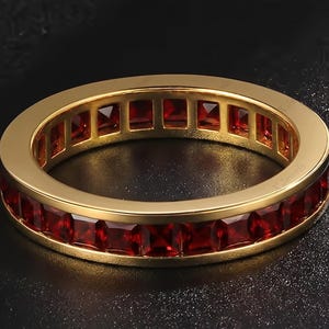 Puede incluir: Un anillo de eternidad en oro con un engaste de canal continuo de piedras preciosas granate de color rojo intenso talladas en forma cuadrada. La banda de metal tiene un acabado pulido y las piedras están dispuestas en una fila uniforme.