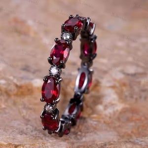 May include: A black metal eternity band featuring alternating oval-cut deep red gemstones and small round clear stones set in a continuous pattern around the entire circumference of the ring, creating a bold and dark aesthetic.