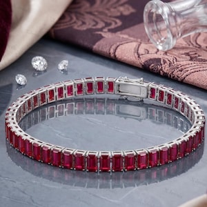 Puede incluir: Una pulsera de tenis en tono plateado con una hilera continua de gemas rectangulares de color rojo intenso engastadas en un marco de metal. La pulsera incluye un cierre de caja seguro y tres piedras pequeñas, sueltas, redondas y blancas.