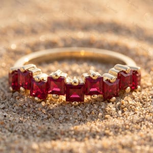 Peut inclure: Une bague en or ornée de sept rubis rouge foncé de taille carrée disposés en rangée. Chaque rubis est maintenu par un sertissage à griffes en or, créant une ligne continue de pierres précieuses sur le dessus de la bague posée sur du sable.