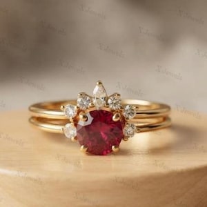 Puede incluir: Un anillo de oro con una gran piedra preciosa redonda de color rojo intenso en el centro, rodeada por un halo de piedras más pequeñas, claras y de corte redondo, con una piedra en forma de pera en la parte superior sobre una banda de oro.
