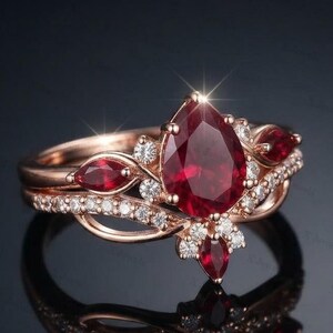 Peut inclure: Une bague en or rose ornée d'une pierre rouge centrale en forme de poire, entourée de petites pierres rouges taille marquise et de pierres blanches rondes. L'anneau présente un design ajouré délicat avec une finition métallique polie.