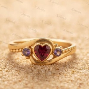 May include: A gold ring featuring a central heart-shaped red gemstone flanked by two smaller round purple gemstones. The band has a smooth, curved design with a beaded texture along the sides, creating a detailed and polished appearance.