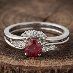 Könnte beinhalten: Ein silberfarbener Ring mit einem zentralen, rund geschliffenen roten Edelstein, der von vier Krappen gehalten wird. Das Band ist mit kleinen, klaren, rund geschliffenen Steinen besetzt, die bogenförmig um den Mittelstein angeordnet sind.