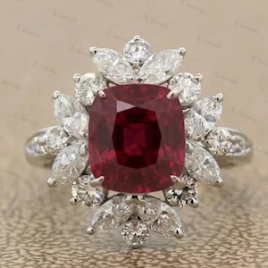 Può includere: Un anello color argento con una grande pietra preziosa rosso scuro a taglio cuscino al centro. La pietra è circondata da un gruppo di pietre bianche trasparenti a taglio marquise e rotondo disposte in un motivo a halo floreale su fascia lucida.