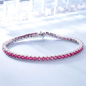 May include: A silver-toned tennis bracelet featuring a continuous row of round, vibrant pink gemstones set in a metallic frame. The bracelet includes a secure clasp mechanism at the end, showcasing a uniform design with sparkling circular stones.