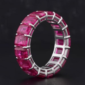 Puede incluir: Un anillo de eternidad completo que presenta una fila continua de gemas de color rosa vibrante con corte esmeralda, engastadas en un marco de metal de tono plateado pulido. El anillo muestra una disposición uniforme de piedras rectangulares sujetas por pequeñas garras.