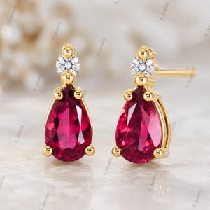 May include: A pair of gold stud earrings featuring a pear-shaped red gemstone at the base and a small round white diamond above it. The earrings are set in yellow gold with a post backing, showcasing a faceted cut on both the red and white stones.