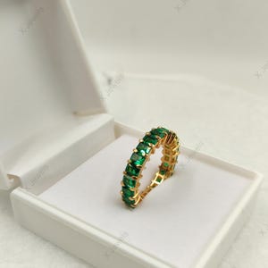 Op de afbeelding: Een goudkleurige eternity ring met smaragdgroene edelstenen. De ring wordt gepresenteerd in een witte sieradendoos. De merknaam "X Jewels" staat op de doos.