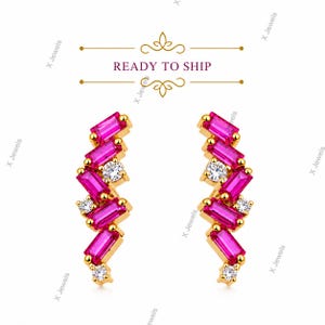 May include: A pair of gold earrings featuring a zigzag arrangement of baguette-cut hot pink stones and round brilliant-cut clear stones. Above the earrings, the text READY TO SHIP is displayed between two decorative gold flourishes.