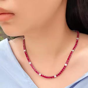 Puede incluir: Un collar de piedras preciosas rojas con piedras blancas de acento. El collar está compuesto por pequeñas gemas rojas redondas, intercaladas con piedras blancas más grandes. El collar se lleva alrededor del cuello, sobre una camisa azul claro.