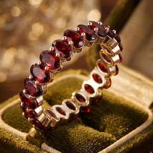 Könnte beinhalten: Ein Eternity-Ring mit einer durchgehenden Reihe von oval geschliffenen, tiefroten Granat-Edelsteinen, die in ein poliertes Goldmetallband gefasst sind. Der Ring ist aufrecht positioniert und zeigt die gleichmäßige Anordnung der facettierten Steine.