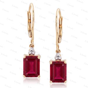 May include: A pair of gold leverback earrings featuring rectangular, emerald-cut red gemstones. Each earring has a small, triangular cluster of three clear, round-cut stones set in gold above the main red gemstone, creating a dangling design.