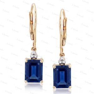 Puede incluir: Un par de pendientes de oro con cierre de palanca, con piedras preciosas azules de corte rectangular suspendidas de un pequeño grupo triangular de tres piedras claras y brillantes. El diseño presenta un acabado de metal dorado pulido.