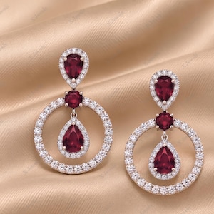 May include: A pair of drop earrings featuring a pear-shaped red gemstone at the top, a small round red gemstone in the center, and a larger pear-shaped red gemstone suspended within a circular frame, all accented with small white stones.