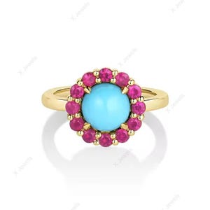 Op de afbeelding: Een gouden ring met een centrale ronde turquoise cabochonsteen, omringd door een halo van kleine, ronde, felroze robijnen. De stenen zijn gezet in een gepolijste gouden band, wat een cirkelvormig, bloemachtig ontwerp met heldere kleuren creëert.