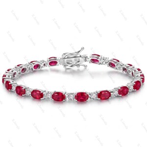 Pode incluir: Uma pulseira de ténis em prata com um padrão repetitivo de rubis vermelhos de corte oval alternados com pequenas pedras redondas transparentes. A pulseira inclui um fecho de caixa metálico seguro com uma marca de contraste 925.