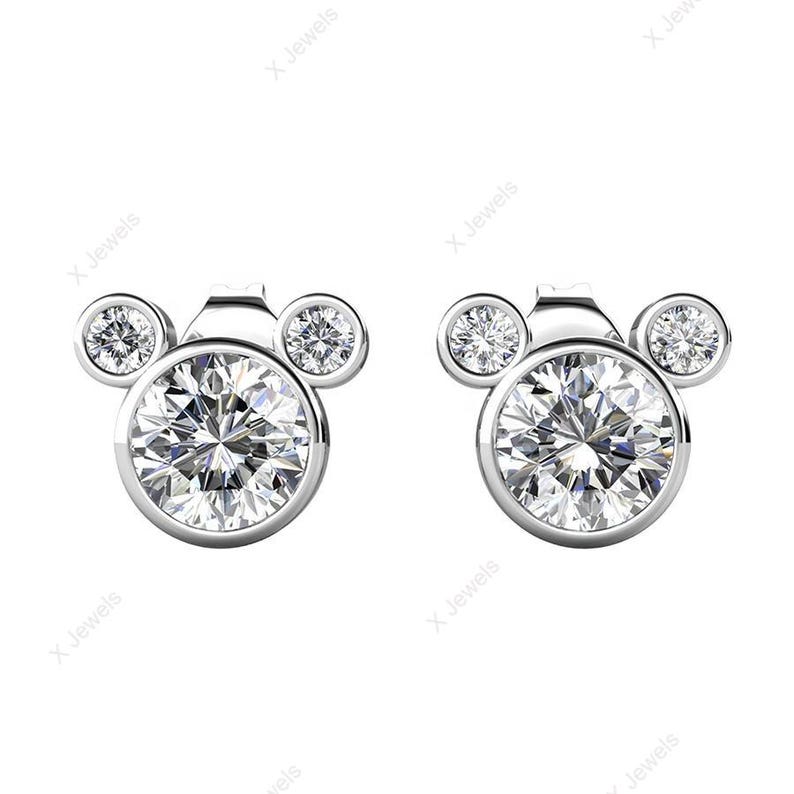 May include: A pair of silver-toned stud earrings. Each earring features a large, round, clear gemstone at the center, with two smaller gemstones above, forming a Mickey Mouse head shape. The gemstones sparkle brightly.