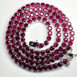 Puede incluir: Un collar plateado con numerosas piedras preciosas redondas de color rojo intenso. Las piedras están muy juntas, creando una línea continua de color. El collar tiene un cierre seguro y una pieza final decorativa, sobre un fondo blanco.