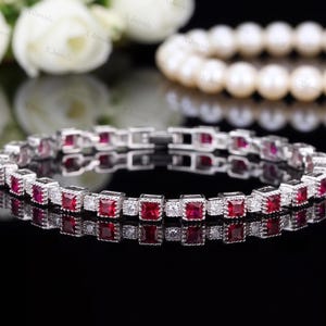 Könnte beinhalten: Ein silberfarbenes Tennisarmband mit abwechselnd quadratisch geschliffenen, tiefroten Edelsteinen und kleinen, klaren weißen Steinen. Das Armband hat eine metallische Gliederkonstruktion mit einem sicheren Verschlussmechanismus.