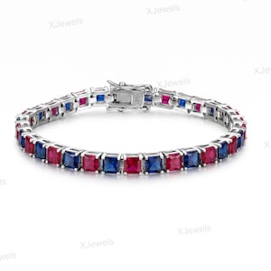 May include: A silver-toned tennis bracelet featuring a repeating pattern of square-cut, vibrant blue and deep red gemstones set in a metallic frame. The piece includes a secure box clasp mechanism and is displayed against a plain white background.