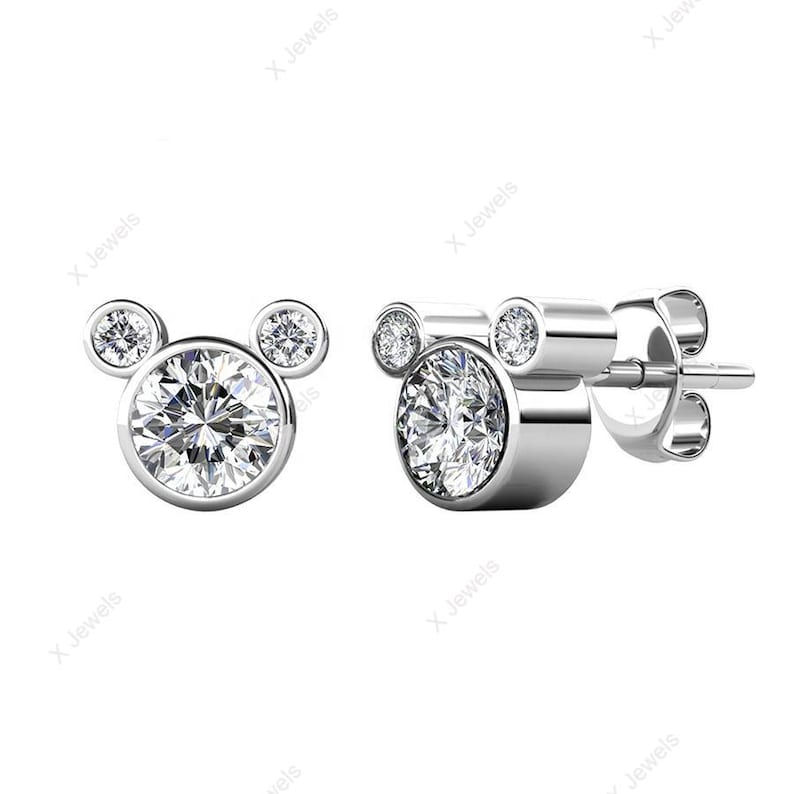 May include: A pair of silver-toned stud earrings. Each earring features a large, round, clear gemstone at the center, with two smaller gemstones above, forming a Mickey Mouse head shape. The gemstones sparkle brightly.