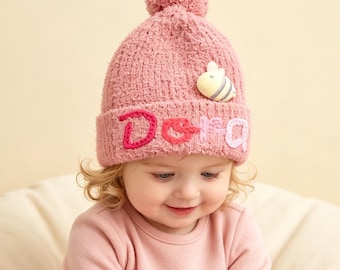 Puede incluir: Un gorro de punto rosa empolvado con un pompón en la parte superior y un borde doblado. El borde muestra el nombre Dora bordado en hilo rosa y blanco, acentuado por una pequeña aplicación tridimensional de abeja amarilla y blanca.
