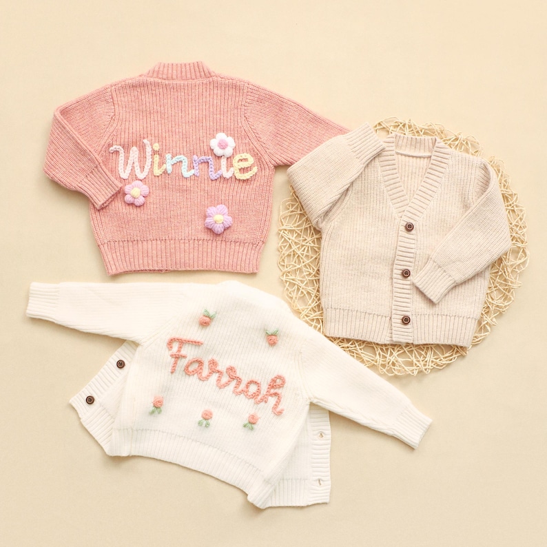 K&ouml;nnte beinhalten: Drei Kinderstrickjacken in einer Studioaufnahme. Ein rosa Pullover mit dem Namen "Winnie" mit Blumen bestickt, eine cremefarbene Strickjacke und ein wei&szlig;er Pullover mit dem Namen "Farrah" mit Blumen bestickt.